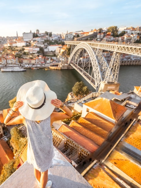 Someone in a white sun hat looks out across the Douro to Porto, the Dom Luís I bridge and the terracotta rooftops